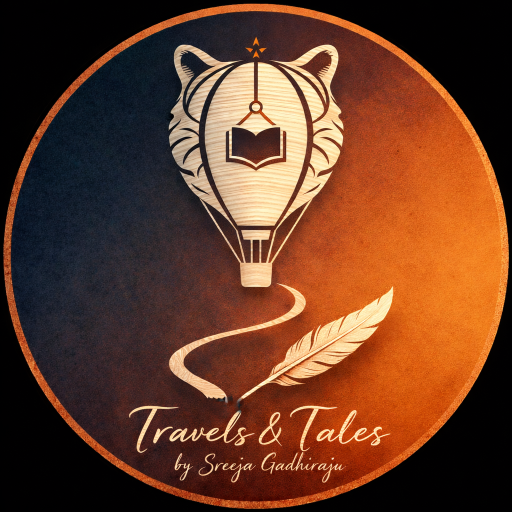 Circular logo with a metallic copper border and a gradient background transitioning from deep navy blue to warm amber. At the center is a hot air balloon subtly shaped like a tiger’s face, featuring faint stripes and ears. Inside the balloon is an open book beneath a small upward arrow symbol. Below, a flowing S-shaped path curves toward a detailed feather. At the bottom, elegant cursive text reads “Travels & Tales” with “by Sreeja Gadhiraju” beneath it in copper lettering.